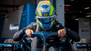 Dino Beganovic gets into his blue DAMS car with his helmet on, hands on the halo. The Ferrari Driver Academy logo is visible on the helmet.