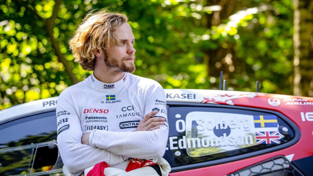 Oliver Solberg stands with his arms crossed, leaning against his Toyota GR Yaris Rally1 in a forest setting during Rally Estonia