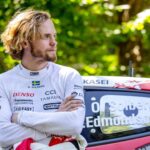 Oliver Solberg stands with his arms crossed, leaning against his Toyota GR Yaris Rally1 in a forest setting during Rally Estonia